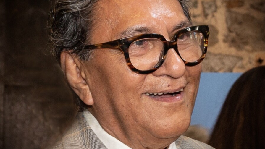 A portrait of Larry Baza from shoulders up. He's wearing thick, tortoise shell glasses, a beige plaid suit with yellow details and a gold tie. Baza is smiling and looking off to the side.