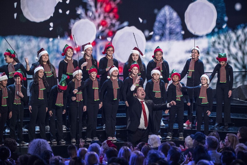 Children wearing all black with green and red striped scarves and Santa hats stand in two tiered rows on a stage as they sing. A conductor stands in the center front, arms outstretched. Behind the children's choir is a wintery backdrop on a LED screen. Audience members can be seen in the front row, watching.