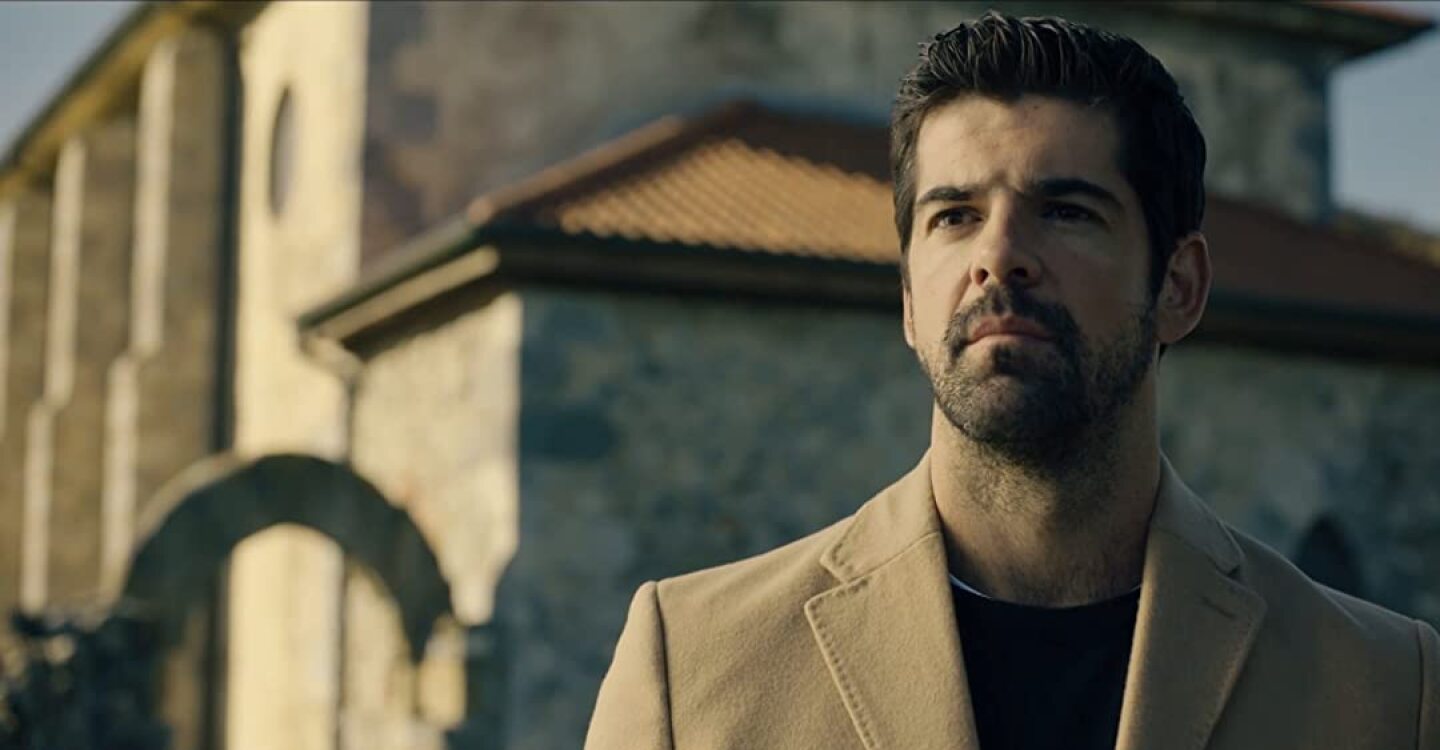 A person wearing a tan blazer looks on with a Spanish-style building behind them.