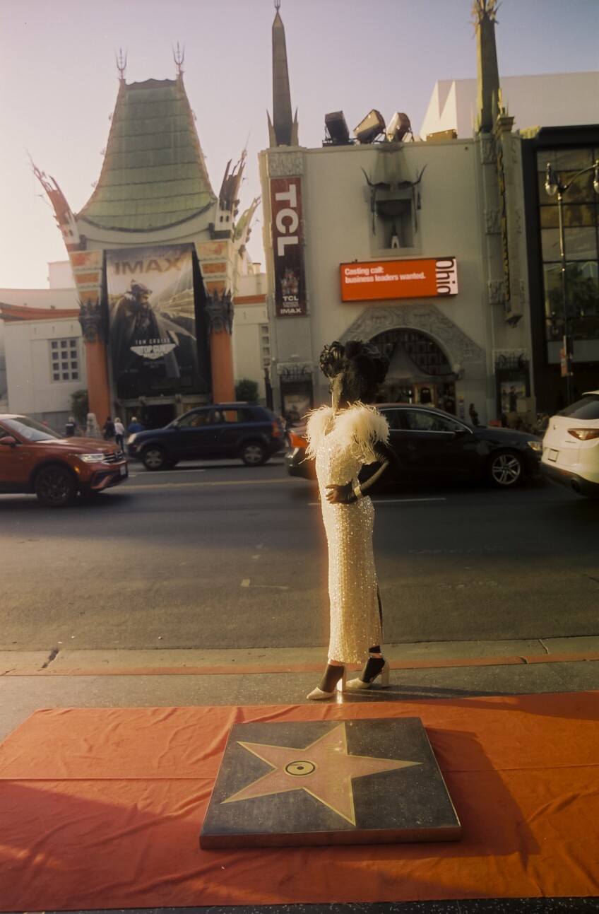 Muxxxe, who is adorned in a black morph suit under a sparkling white dress, curled wig and bedazzled jewelry all meant to recall old Hollywood glamor, stands behind their Walk of Fame star recreation. The slab sits on top of a red carpet. Behind Muxxxe is the Grauman's Chinese Theater and cars bustling down the street.