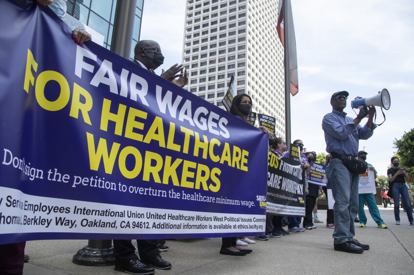 Masked demonstrators carry a banner that reads "FAIR WAGES FOR HEALTHCARE WORKERS"