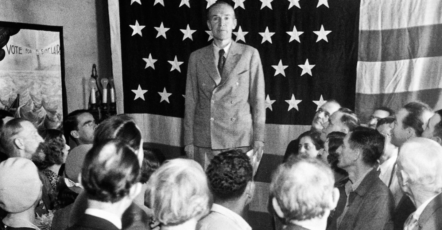 Upton Sinclair stands among seated people with an American flag tapestry hung up behind him.