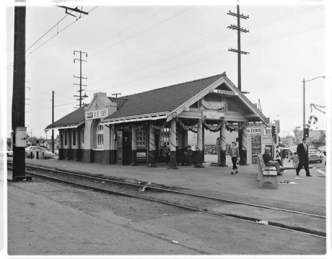 A black and white photo of the Lynwood Depot, with columns lined with Christmas decorations. The depot stands next to a set of train tracks. Passengers and pedestrians walk in the sidewalks near the depot while others sit on benches, waiting.