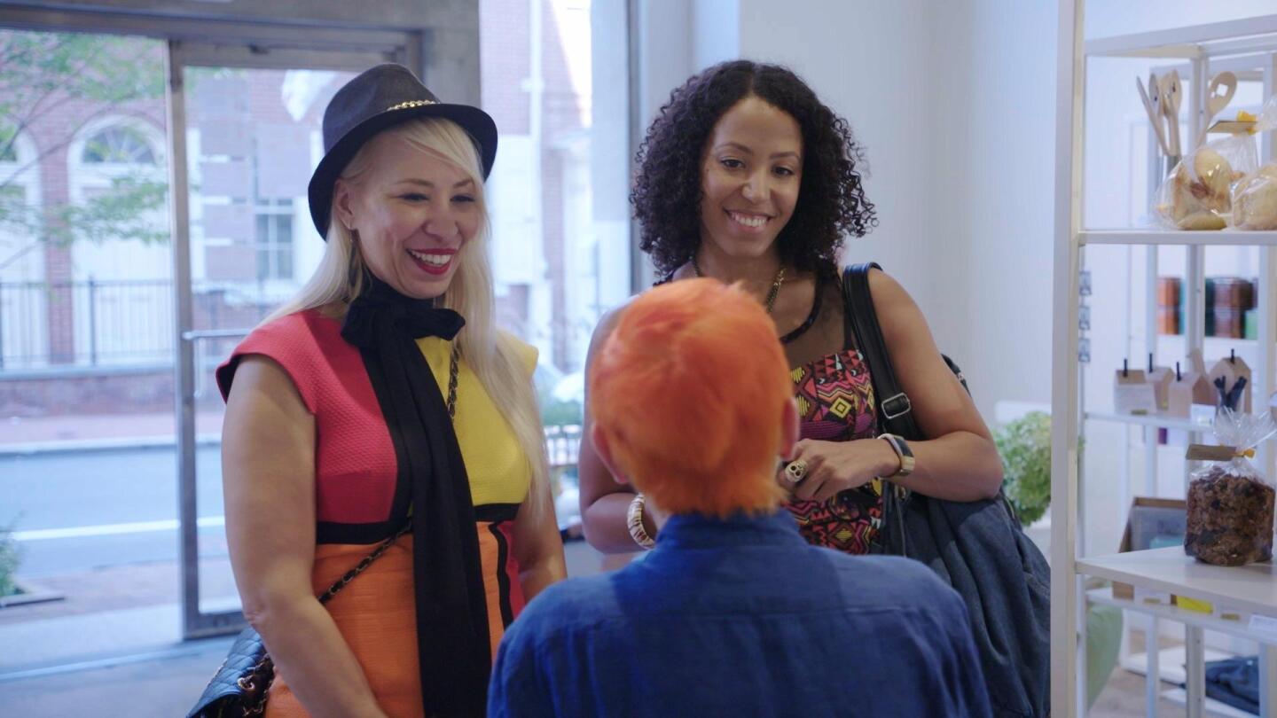 Two smiling people face an orange-haired person inside a shop.