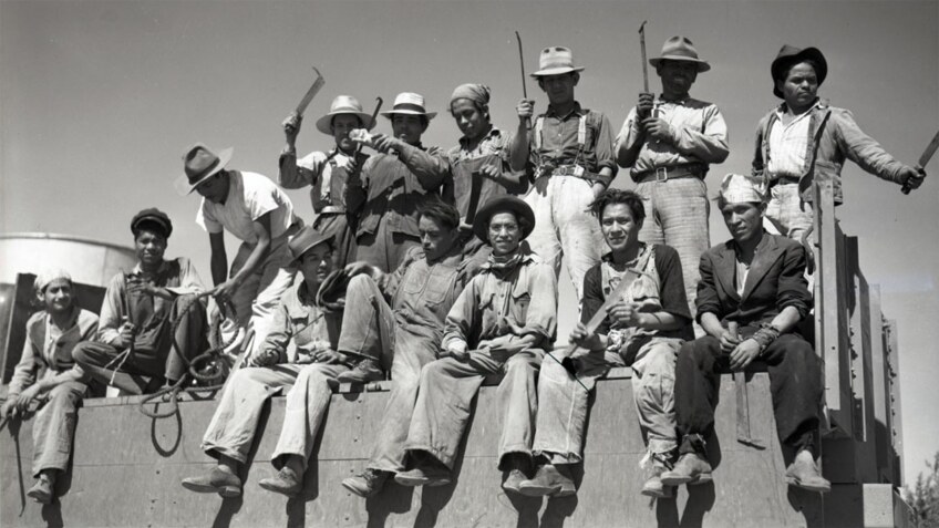 A photo of braceros sitting on the back of a flatbed truck holding hoes, 1942.