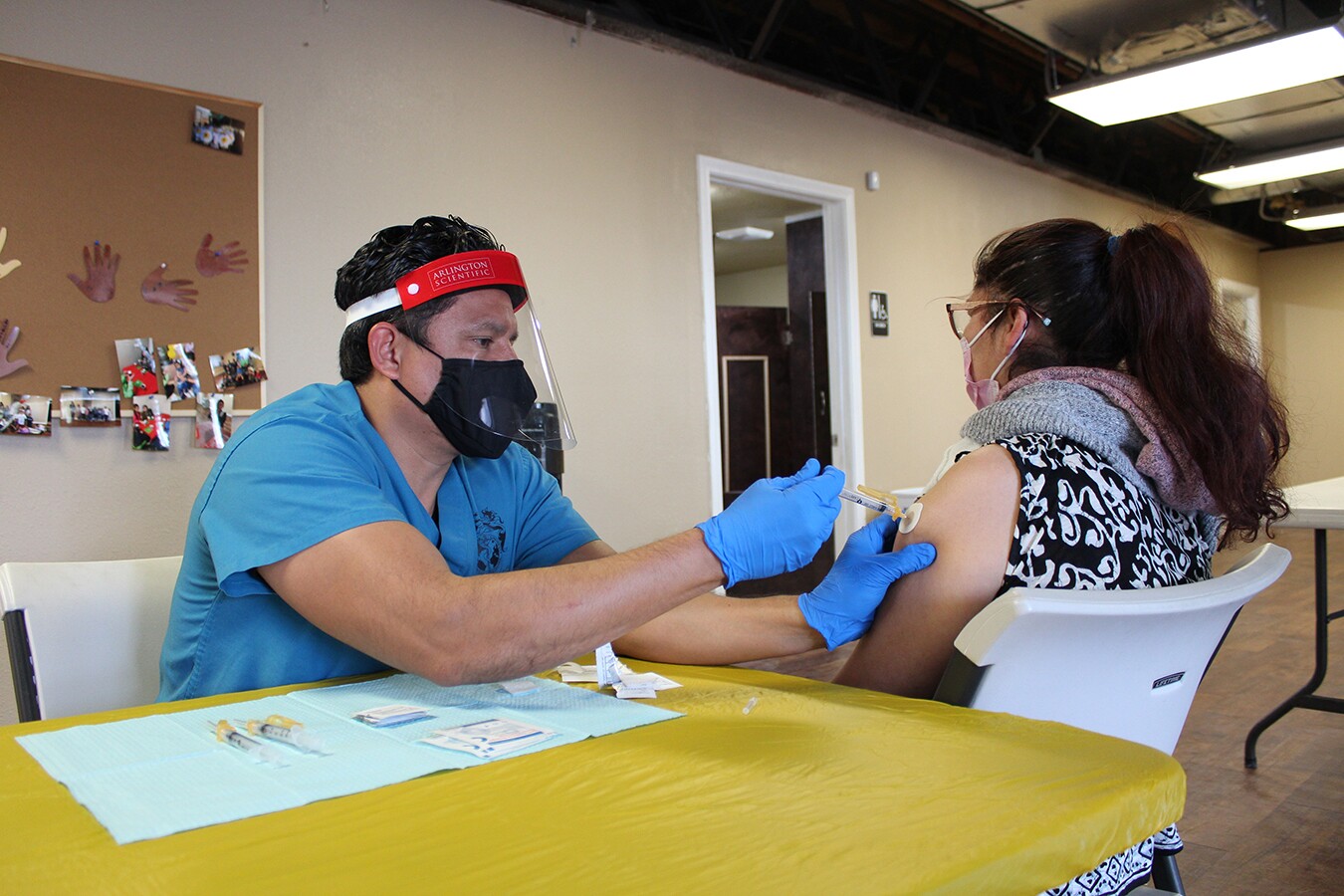 A woman receives her COVID vaccine by a yellow table. The shot is being administered by a man in teal blue scrubs.