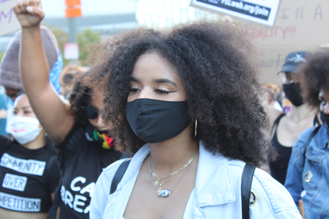 Catherine Estrada in a black mask with people marching with signs behind her.