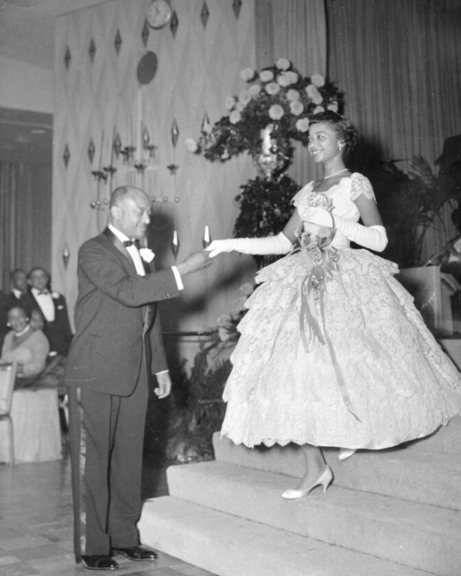 A young African American debutantes walks down the stairs holding her father's hand.