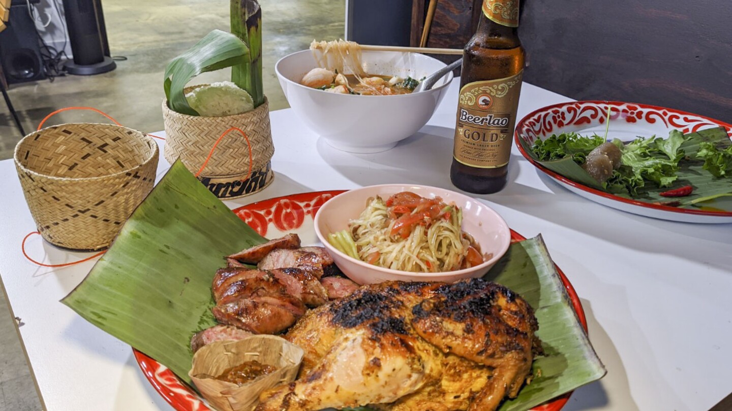 A table is prepared with various Lao dishes. The plate in the foreground is a papaya salad with Lao sausage and chicken. Behind it is a bowl of sukiyaki. To the left of the papaya salad is sticky rice nestled in a small woven basket and to the right is a plate of sakoo yut sai or tapioca dumplings. A bottle of beer also sits on the table.