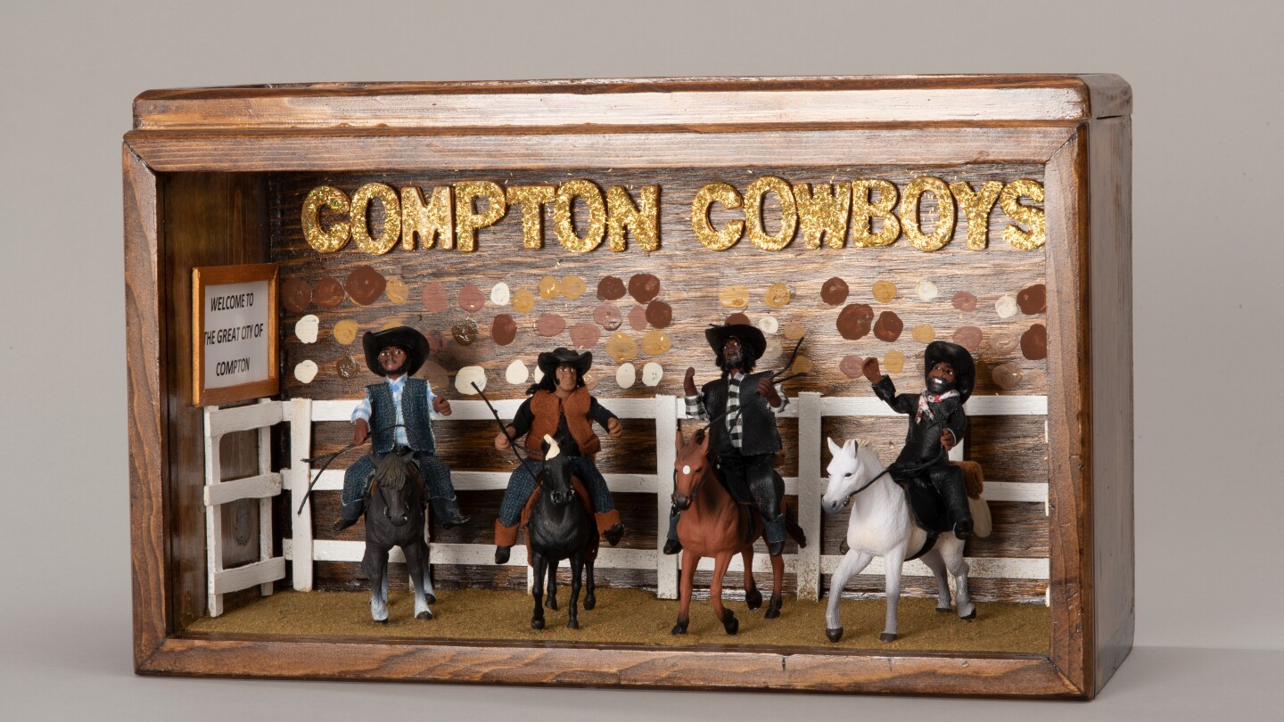 A wooden shadowbox diorama with four miniature Black men on horseback. They're all wearing cowboy hats and cowboy attire. Behind them is a miniature white fence with various skin-toned dots painted onto the back of the shadowbox, implying a crowd of people beyond. Gold, sparkly block letters read, "Compton Cowboys."