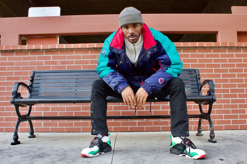 A young person wearing a multicolored jacket stares ahead while sitting on a bench. 