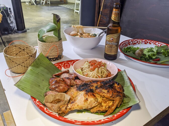 A table is prepared with various Lao dishes. The plate in the foreground is a papaya salad with Lao sausage and chicken. Behind it is a bowl of sukiyaki. To the left of the papaya salad is sticky rice nestled in a small woven basket and to the right is a plate of sakoo yut sai or tapioca dumplings. A bottle of beer also sits on the table.