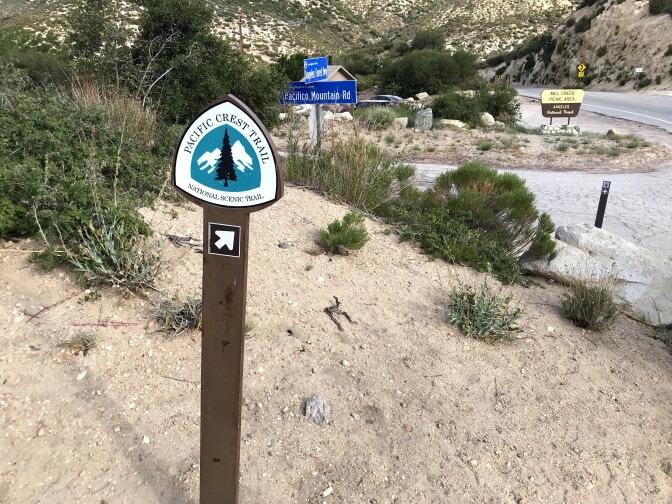 A Pacific Crest Trail signpost can be found on a hillside with the road sign Pacifico Mountain Road sign in the background.