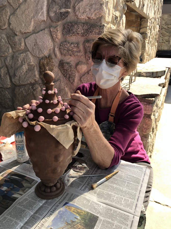 An elder woman with sunglasses wears a face mask while fine tuning her ceramic creations.