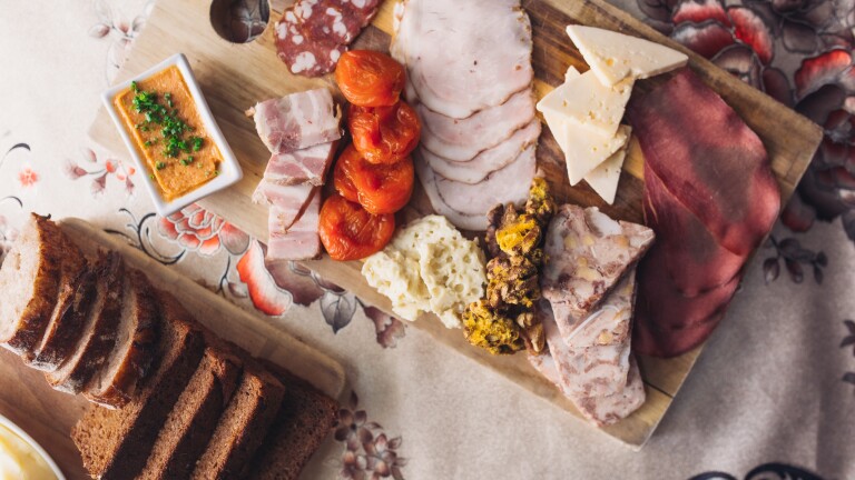 Slices of bread, cheeses and cured meats are arranged on wooden board that sits atop an ornate and decorative fabric tablecloth. 
