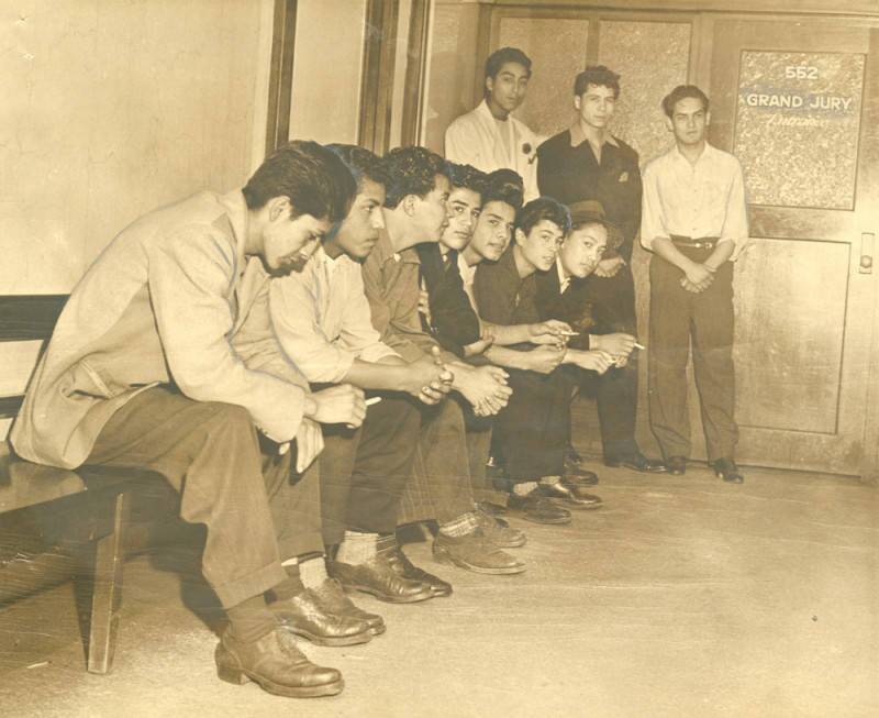 A sepia-tone photo shows a row of young Latino men sitting against a wall on a bench while three others stand against a wall where a door reads "GRAND JURY"