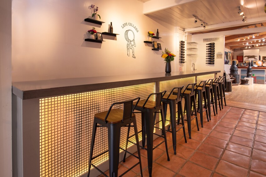 An indoor wine tasting room. Bar stool chairs are lined up against a counter with internal lightning. Behind the counter are shelves with flower vases and wine bottles. On the wall, reads "Lepe Cellars" over a graphic of a man holding a wine barrel on his back. 