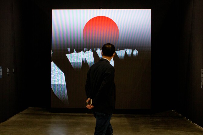 A man in a suit with his hands behind his back looks on to a digital art piece on a large LED screen mounted on a black gallery wall. The digital art piece features a large red dot resembling a setting sun with floating white