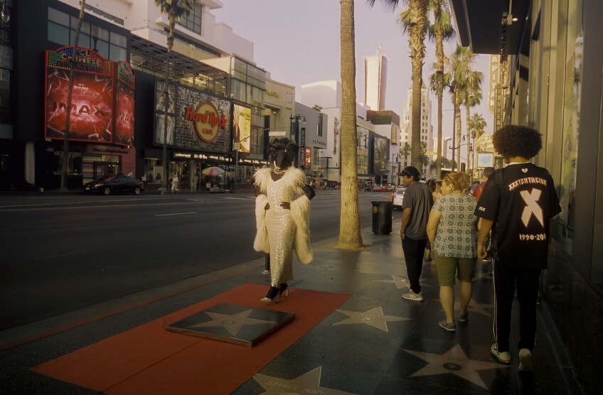 Muxxxe, who is adorned in a sparkling white dress, curled wig and bedazzled jewelry stands on a red carpet laid out on the Hollywood Walk of Fame. On the carpet in front of them is a Hollywood star recreation. Pedestrians look on as they walk by.