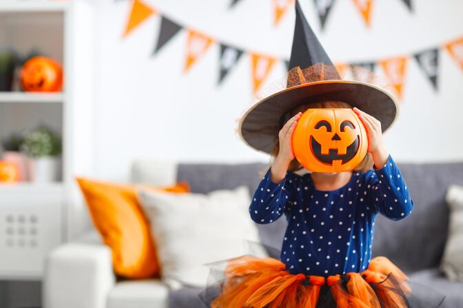 A little girl in a witch hat holds a plastic jack o' lantern over her face.