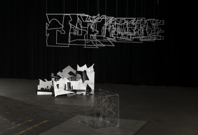 Three sculpture works stand in frame. Two are on the floor and resemble sharp, angular, abstract shapes. While one hangs from the ceiling.