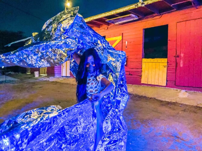 A performer wearing a mask holds a foil object around her. The performance is part of at "Firefly Nights," an in-person COVID-safe experience that falls within the "Rio Reveals" series.