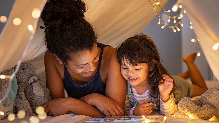 A mother and child watch a tablet together under a sheet decorated with small lights. iStock