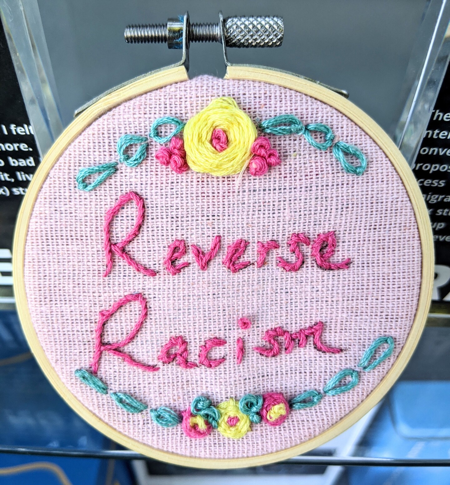 A small embroidery hoop with a pastel pink fabric stretched across embroidered with the words "Reverse Racism" in hot pink thread. The words are framed with a floral embroidery design featuring yellow flowers, pink details and green leaves.