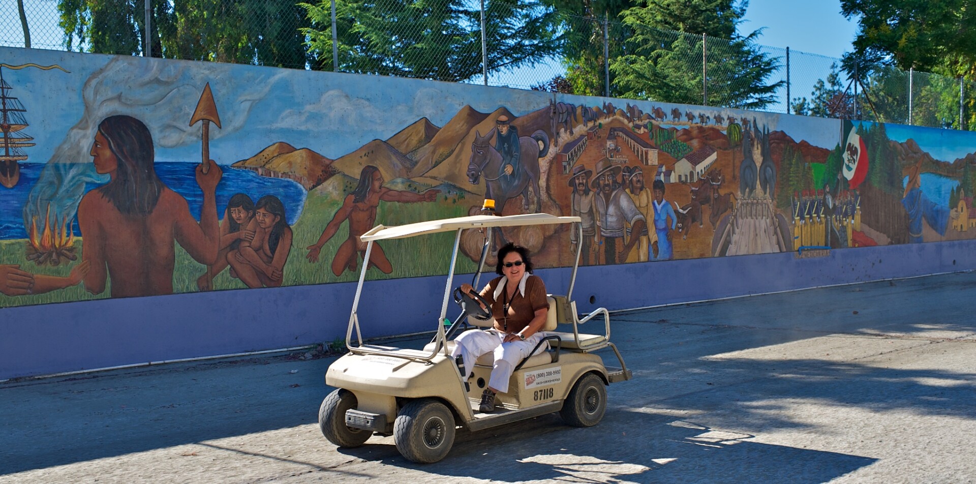 A woman in a small golf cart drives along beside a long mural in a riverbed.