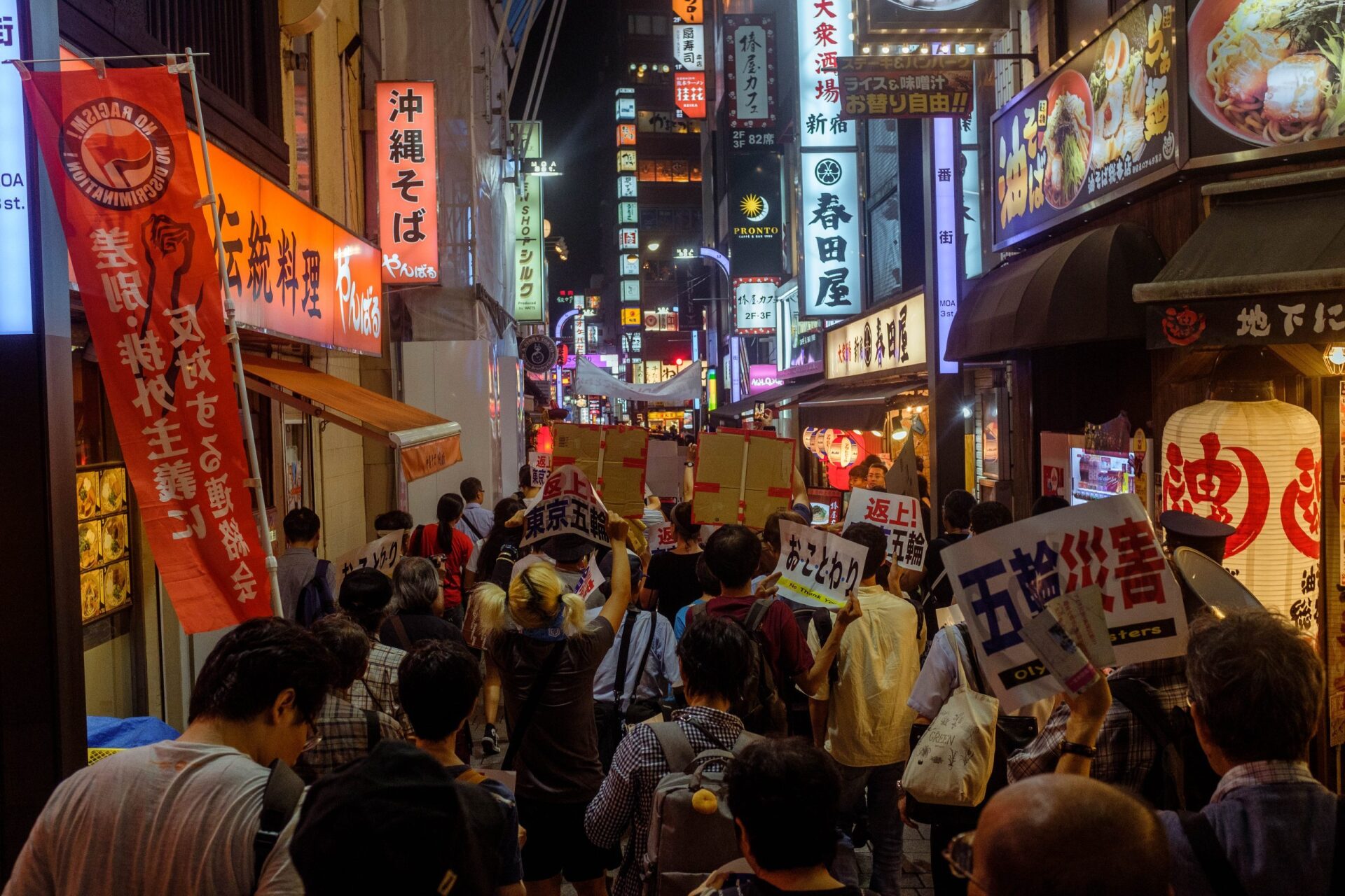 A large crowd with signs in Japanese marches through a brightly-lit alleyway in Shinjuku, Japan.