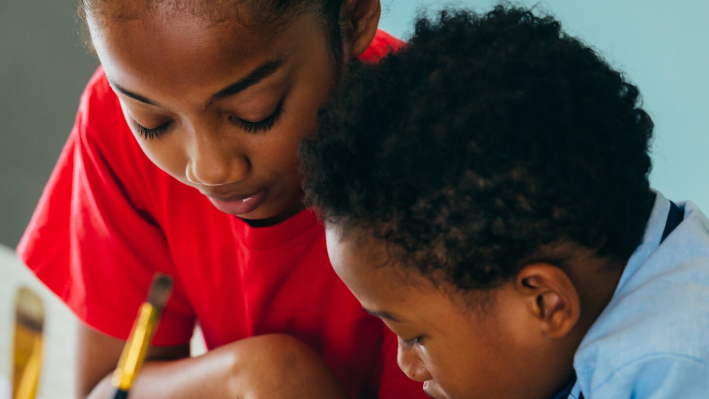 Two Black children lean into something they are working on on a table with paintbrushes and crayons.