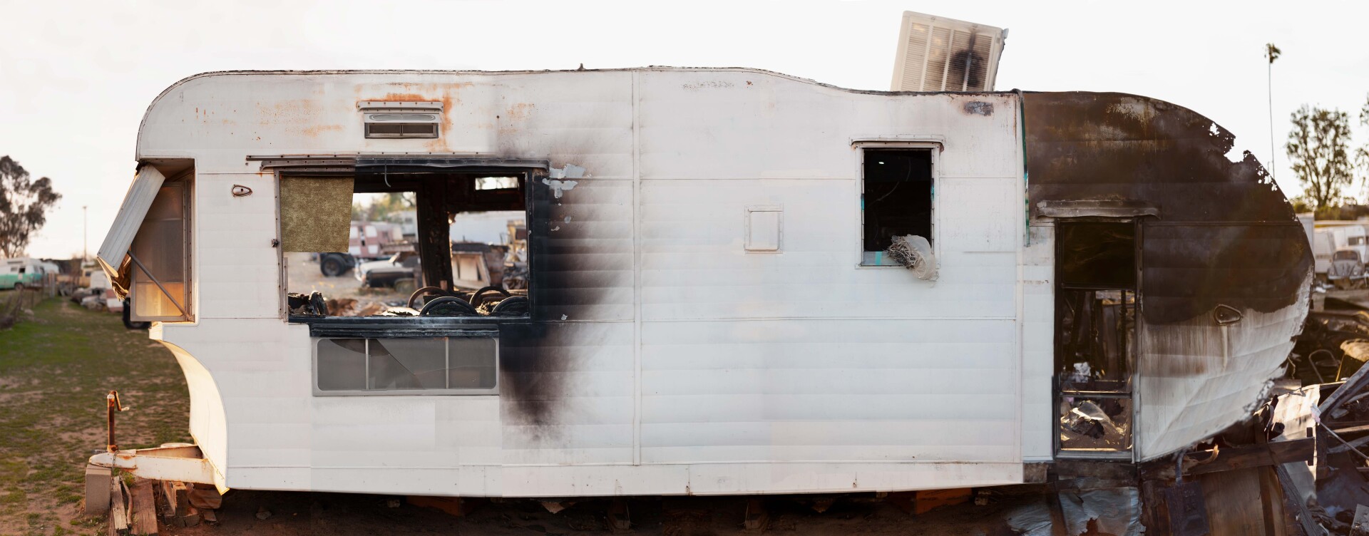A deteriorated white trailer with dilapidated edges and singed edges, black from fire. The doors and windows are ripped out of the trailer, revealing a messy interior.