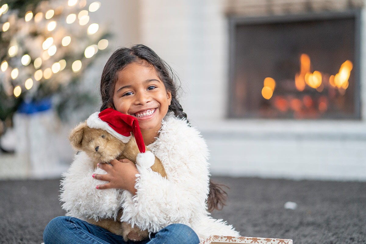 A small child sits in front of a fireplace and a tree with twinkly lights. She is holding a teddy bear and smiling. iStock