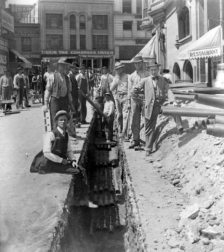 A black and white photo of men gathered around a trench in the road. Pipes run across the bottom of the trench.