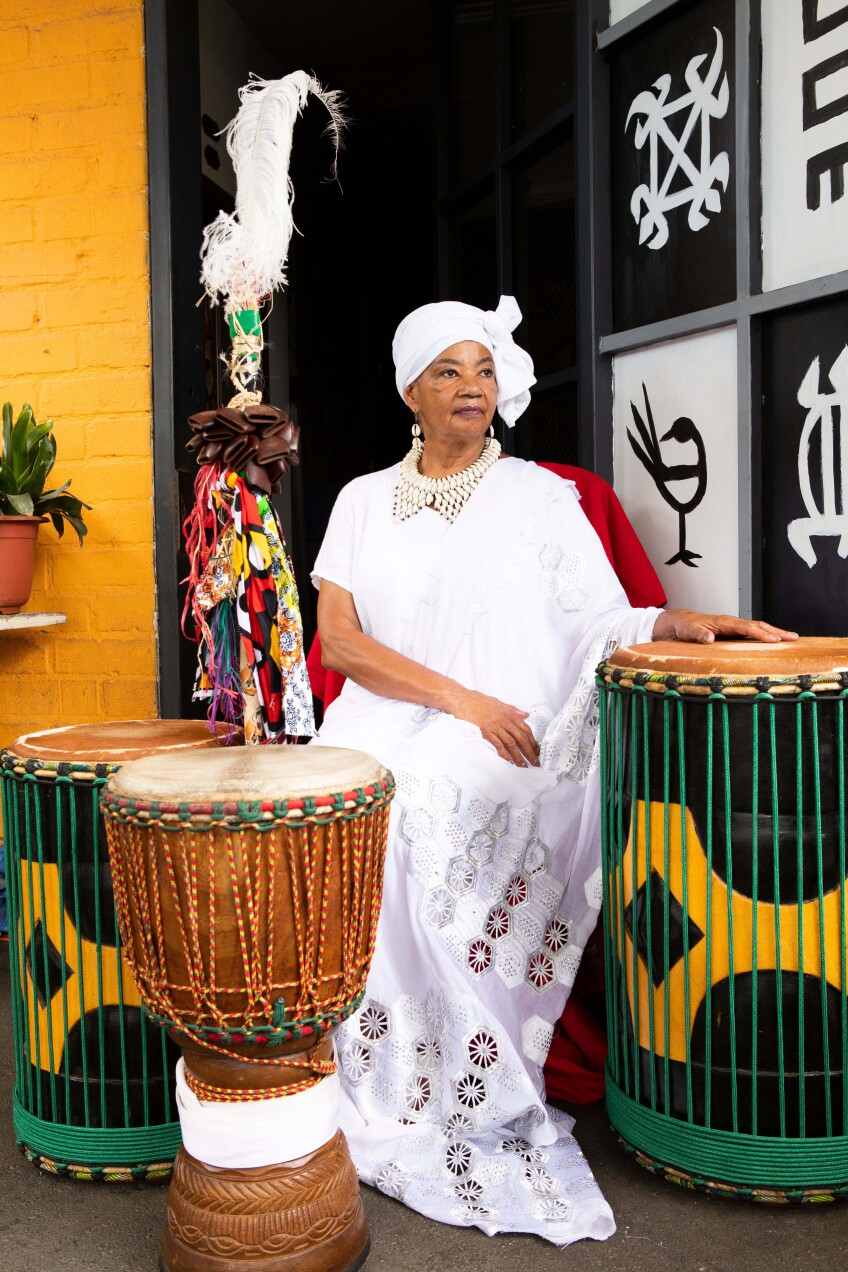 A woman in West African dress (including a long, white garment and matching headscarf) sits among West African drums adorned with colorful rope.