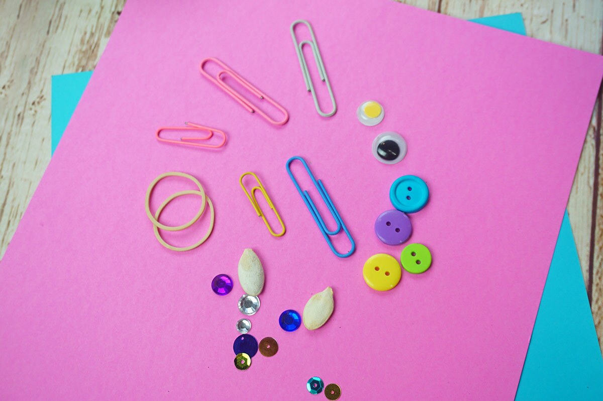 A pink piece of paper covered with assorted small items including google eyes, rubber bands, paper clips, buttons, seeds and sequins.
