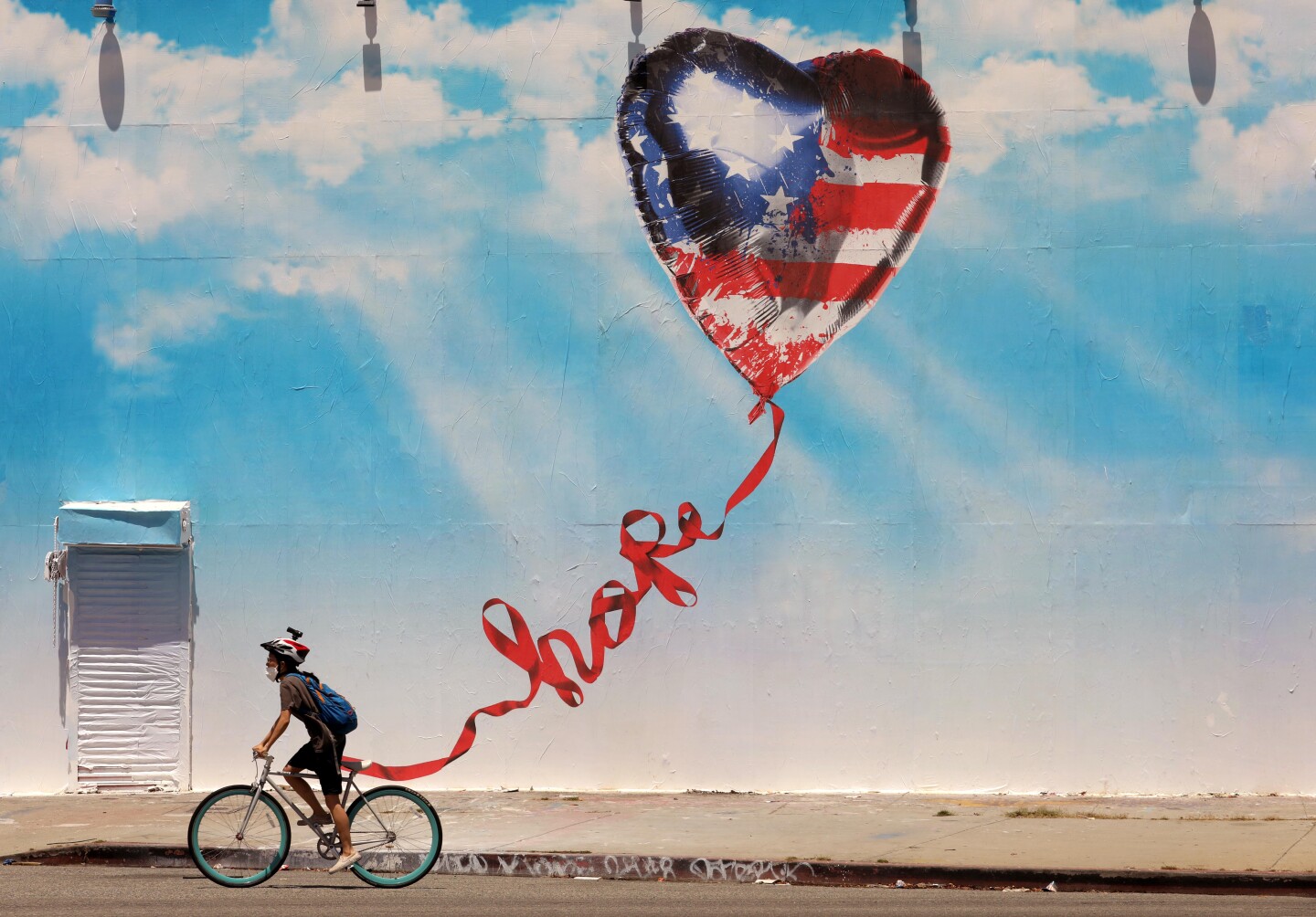 A bicyclist wearing a face mask rides past a mural depicting a heart-shaped balloon patterned like the American flag flying in a blue sky. The balloon's string is twisted to read "hope."