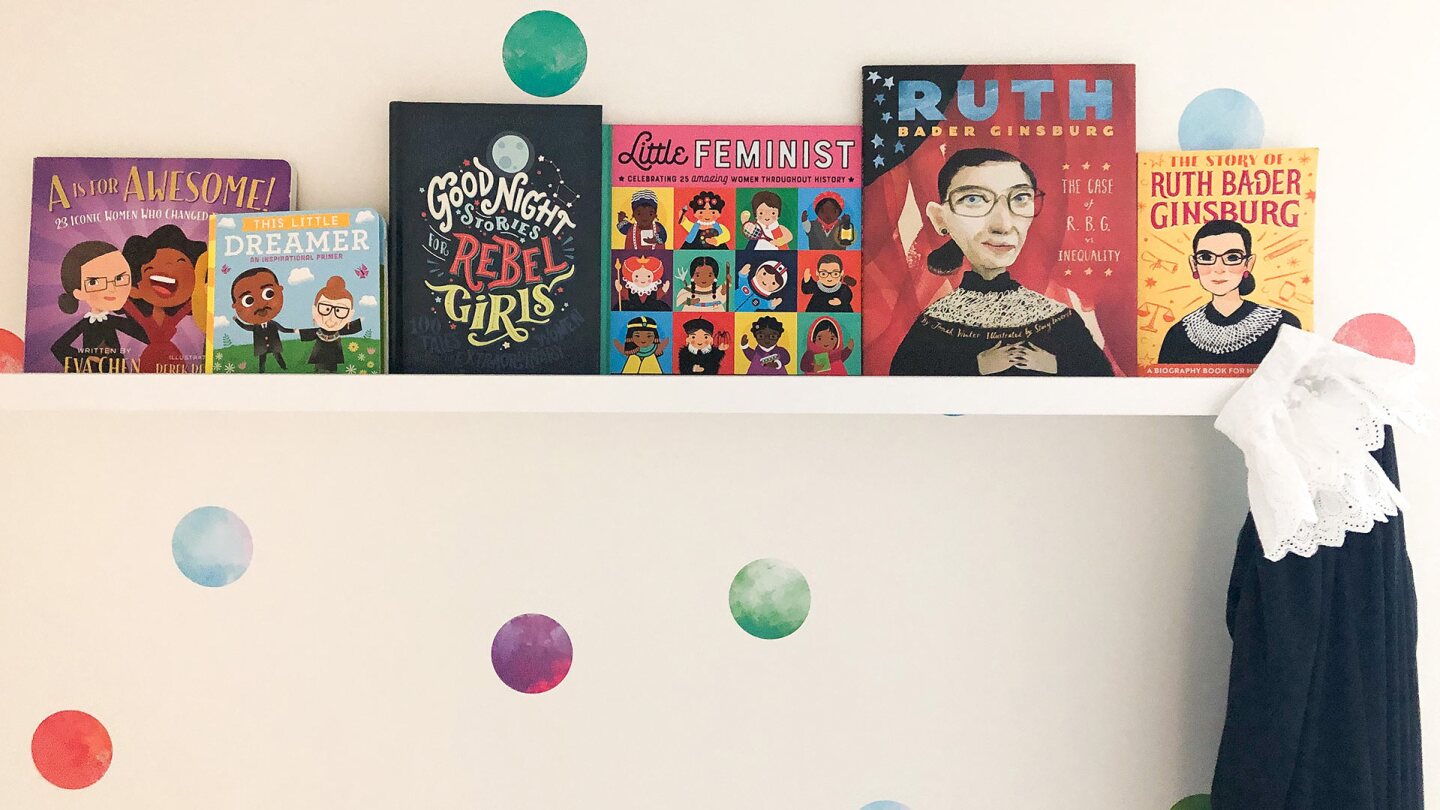 A bookshelf on a wall is filled with 6 books featuring historic women and/or Justice Ruth Bader Ginsburg. A black clock with a white lace collar hangs from it.