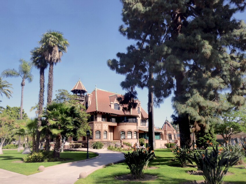 The Doheny mansion, an red-orange brick building of French Chateauesque style architecture, mixed with Gothic, Moorish and California Mission elements stands among a grove of various trees. A paved pathway winds up to the mansion. Above the mansion is a bright blue sky.  