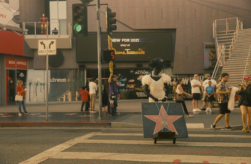 Muxxxe, who is adorned in a sparkling white dress, curled wig and bedazzled jewelry over a morph suit, pushes a Hollywood Walk of Fame star recreation on a dolly as they cross the street. The star recreation reads, "Muxxxe."