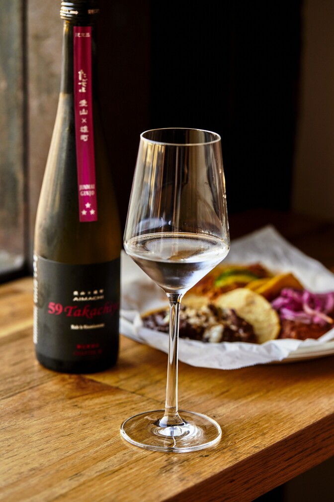 A bottle of unpasteurized nama in a wine glass is set in front of street tacos.