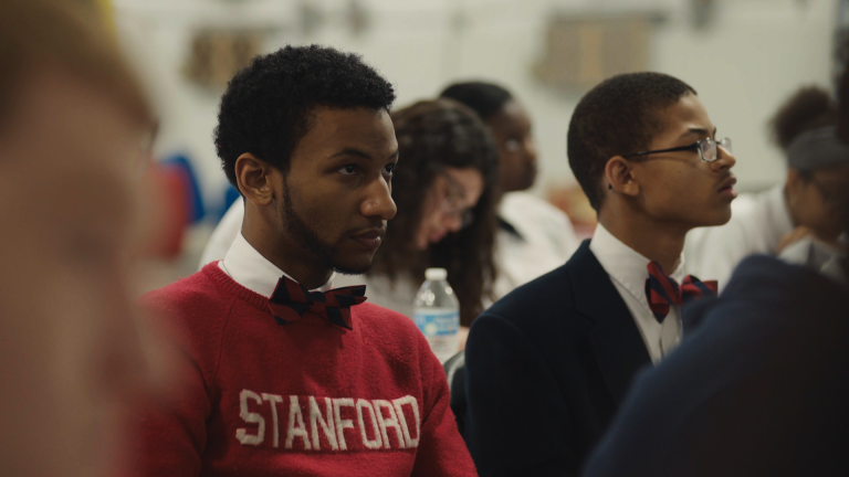Two people wearing bow ties sitting among a crowd look intently in front of them.