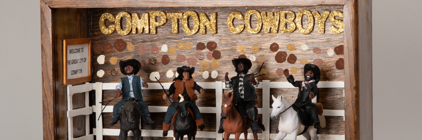 A wooden shadowbox diorama with four miniature Black men on horseback. They're all wearing cowboy hats and cowboy attire. Behind them is a miniature white fence with various skin-toned dots painted onto the back of the shadowbox, implying a crowd of people beyond. Gold, sparkly block letters read, "Compton Cowboys."