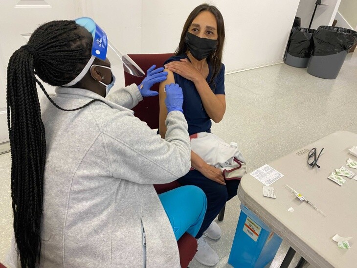 Lina Vargas holds her sleeve up as another woman administers the COVID-19 vaccine into Vargas' right arm.