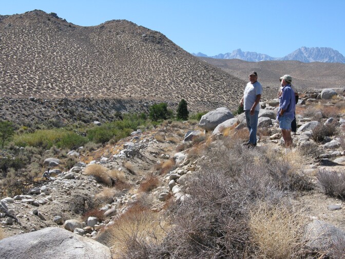 Three men stand at mid-distance at the edge of a dry ditch bed