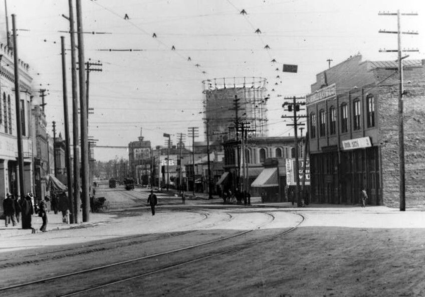 A black and white photo of an unpaved, dirt street. Buildings line the street on either side and electrical lines line the sides of the road, propped up by poles. A gas holder, which resembles a silo, looms over the scene in the distance. Pedestrians walk on the sidewalk, near the buildings.