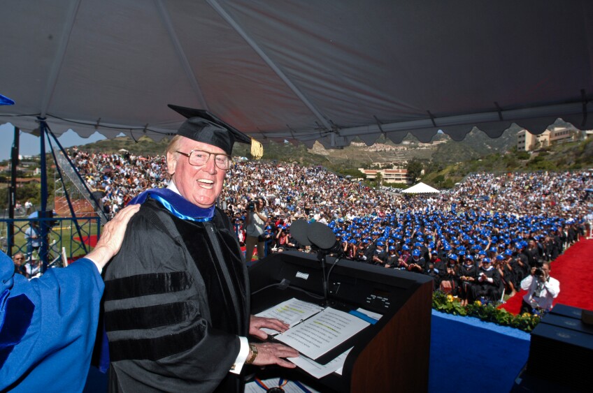 Vin Scully wearing a graduation toga addressing a crowd wearing Dodgers hats with the special embroider in the back.