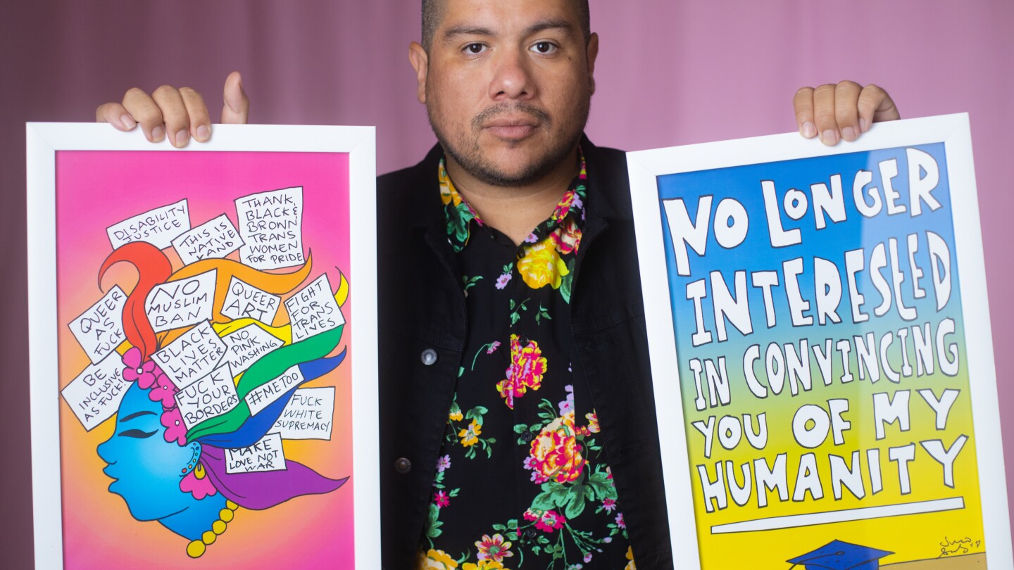 Julio Salgado is wearing a floral print shirt and a black jacket while holding up two pieces of his art on each hand. The artwork on his left features the side profile of a woman with multicolored hair and statements like, "Black Lives Matter," "#MeToo," "Make Love Not War," and "Thank Black and Brown Trans Women for Pride." The artwork on the right reads, "No Longer Interested in Convincing You of My Humanity," with a graduation cap at the bottom. Salgado is standing in front of a pink background.