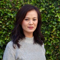 A headshot of Thuy Vo Dang. She has medium-length black hair and is wearing a gray knit sweater. She's standing in front of a green leaf background.