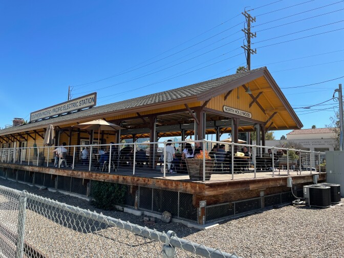 A train depot with yellow wood horizontal paneling and dark brown trim stands against a bright blue sky. A large sign is posted on the roof and reads, "Southern Pacific - Pacific Electric Station." At the back of the depot, where passengers would typically wait for trains, is outdoor seating for a café. Patrons sit in clusters around tables.